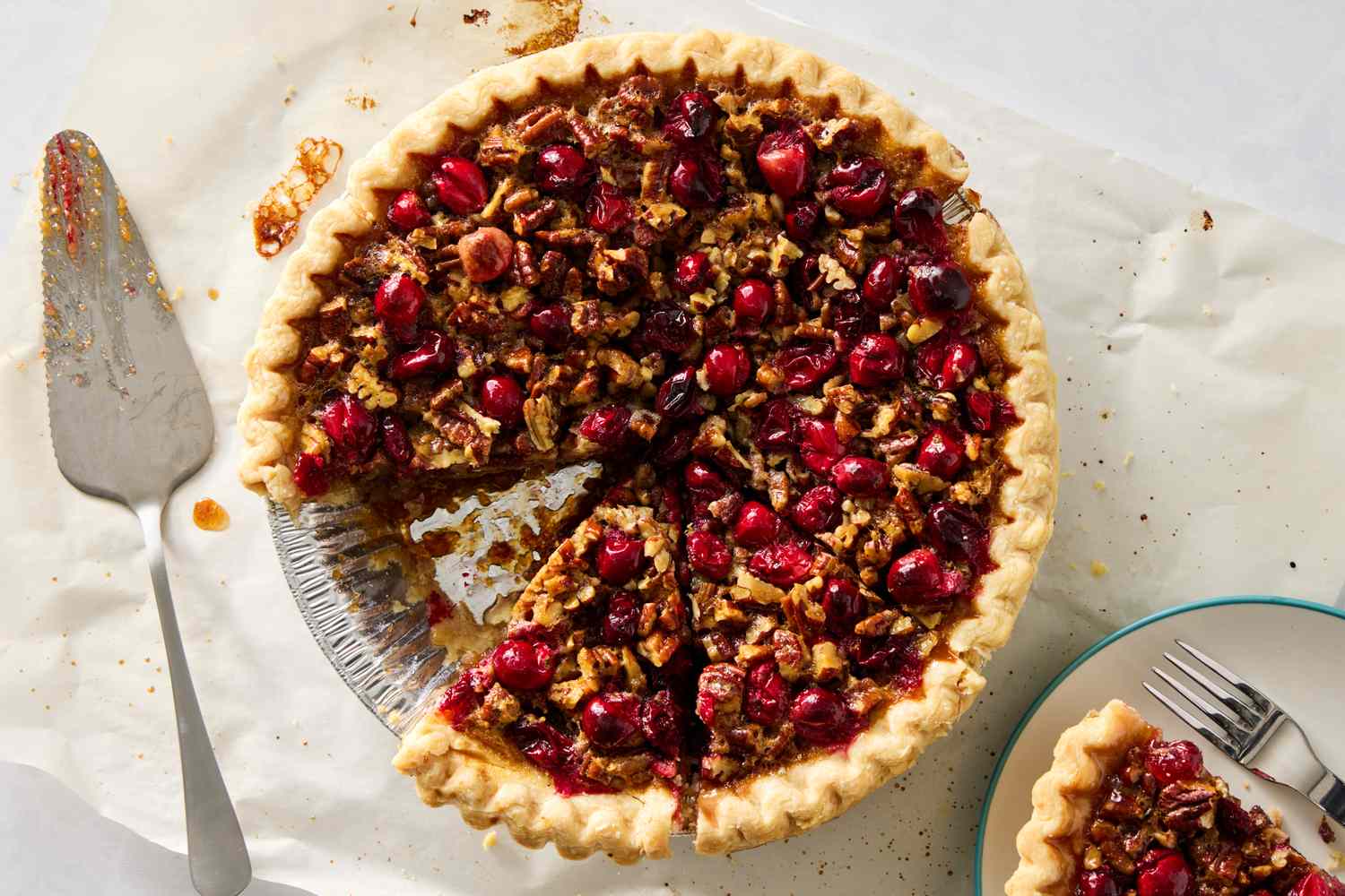 A cranberry pecan pie with a slice removed served on a plate nearby with a fork shown on parchment paper with a pie server