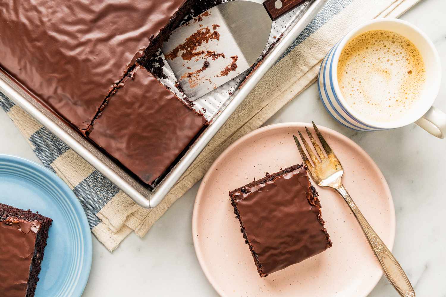 Slices of chocolate depression cake on small plates at a table setting with a baking pan with leftover cake, a cup of coffee, and a kitchen towel