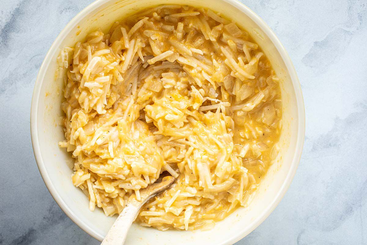 A large white mixing bowl on a marble countertop. Shredded potatoes and cheese mixed together for Hash Brown Potato Casserole.