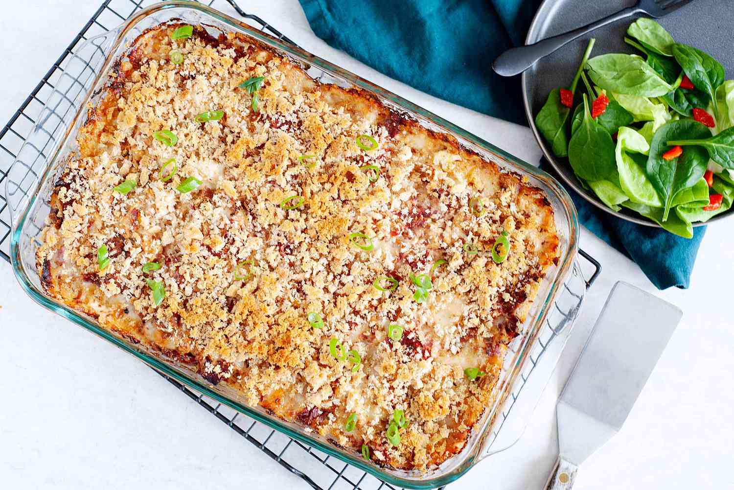A golden brown baked reuben casserole is cooling on a rack. A metal spatula is in the lower right hand corner. A matte grey plate has a side salad and black fork in the upper right. A dark teal linen is under the plate.