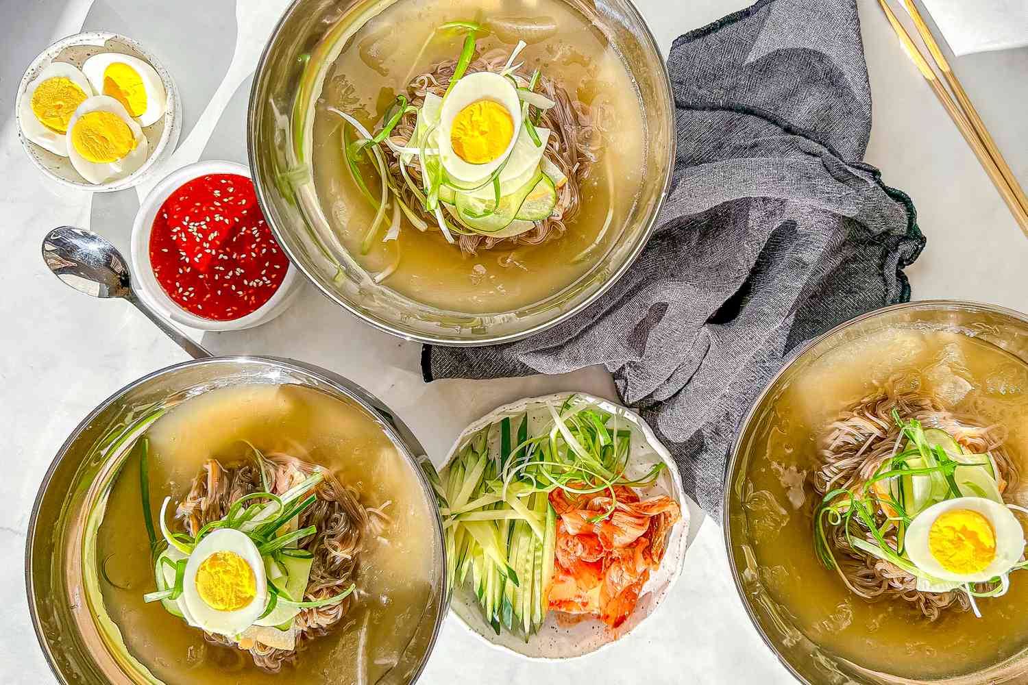 Three Bowls of Mul Naengmyeon Surrounded By Bowls of Toppings (One Bowl With Sliced Cucumbers, Kimchi, and Julienned Green Onions, Another With Spicy Gochujang Sauce, and a Bowl of Halved Hard Boiled Eggs), a Grey Kitchen Towel, and Utensils (a Spoon and Pair of Chopsticks)
