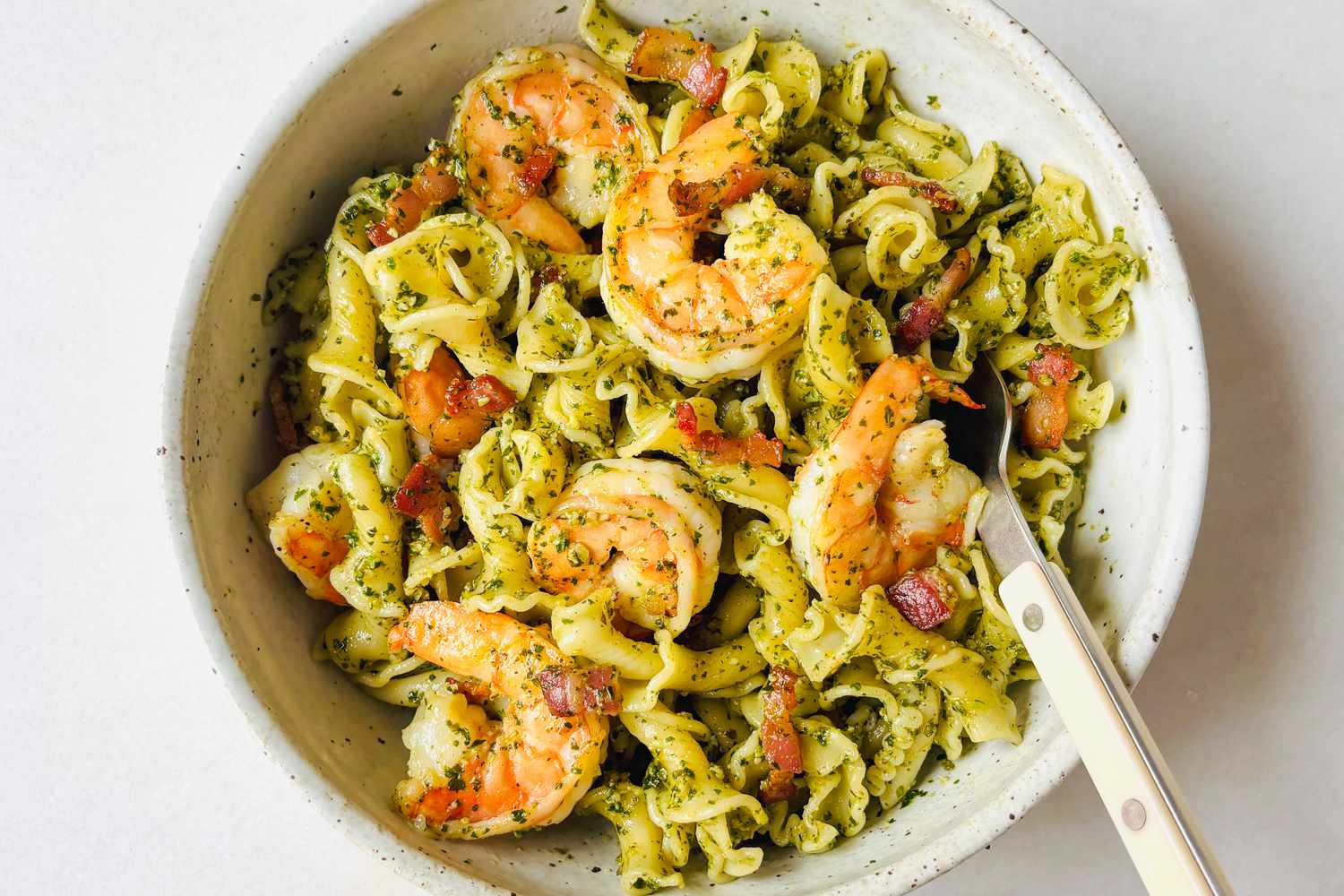 A bowl of pasta topped with shrimp and a green sauce, garnished with small bacon pieces and served with a fork