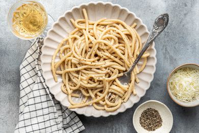 Plate of Brown Butter Cacio e Pepe with Some Noodles Wrapped around a Fork, and Next to It, a Kitchen Towel, a Glass of Wine, and Two Small Bowls (One with Cracked Black Pepper and Another with Shredded Parmesan) 