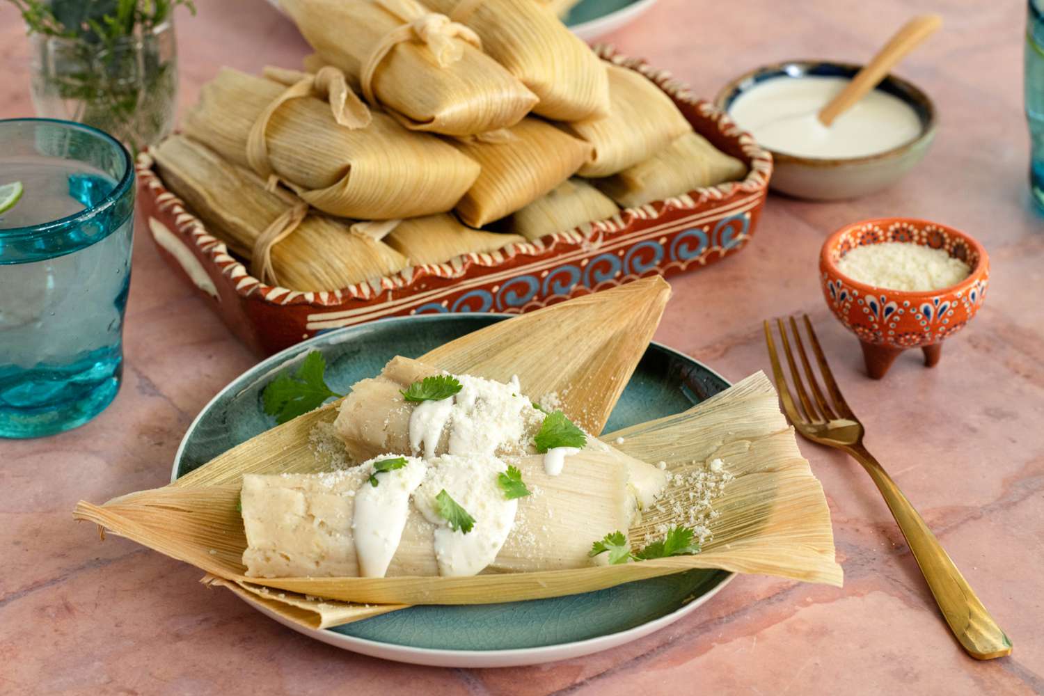 A Plate of Chicken Tamales with Chile Verde Topped with Crema, Cotija Cheese, and Some Cilantro at a Table Setting with a Platter of More Tamales, Utensils, a Small Bowl of Cotija Cheese, a Small Bowl of Crema, a Glass of Water, and a Small Vase
