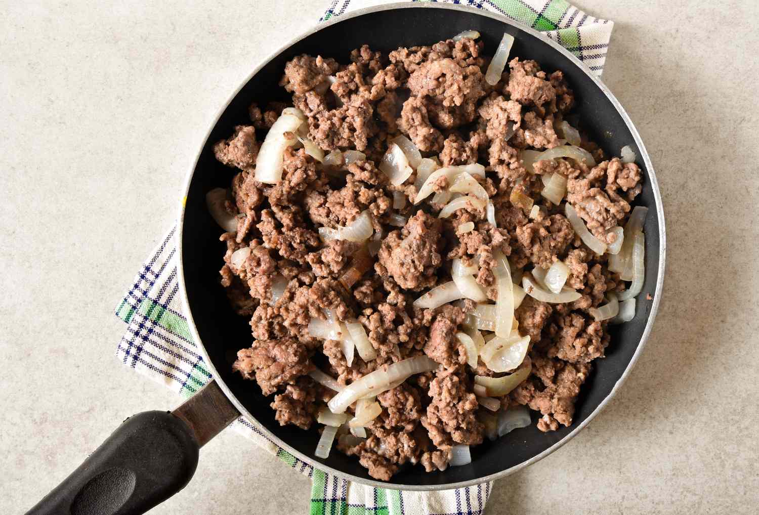 Cooked ground beef with sliced onions in a skillet set on a checkered cloth background
