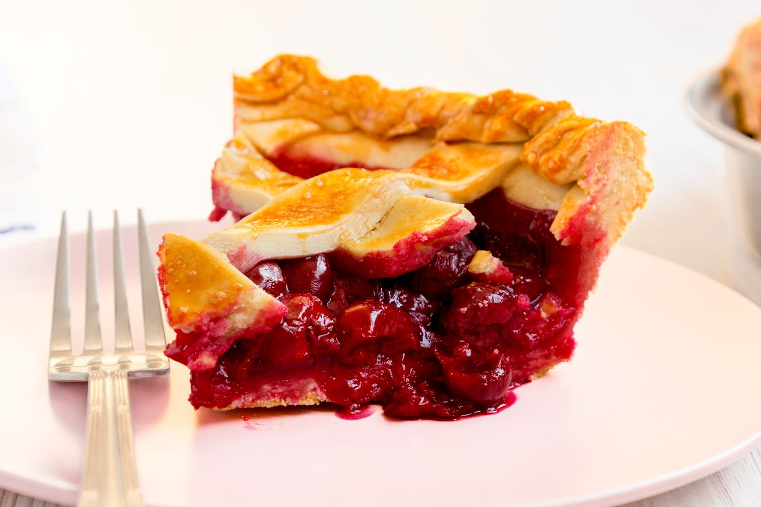 A slice of cherry pie on a pink plate with a fork beside it the filling is visible and surrounded by a flaky crust