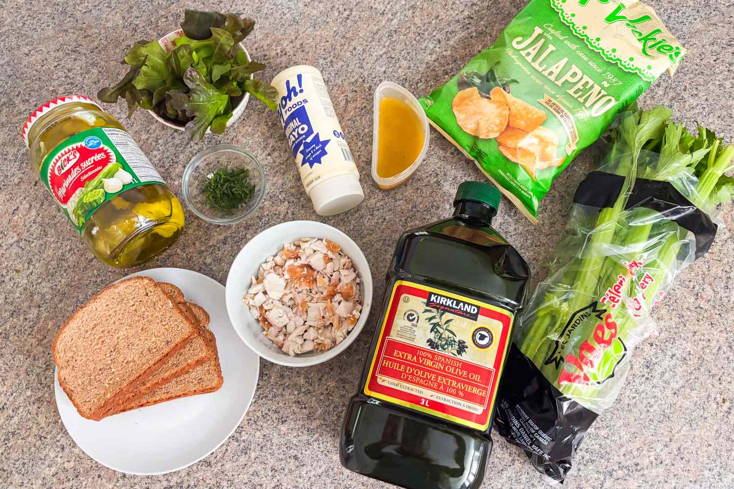 Ingredients for a recipe layout on a counter including bread greens pickles oil sauce and chopped chicken