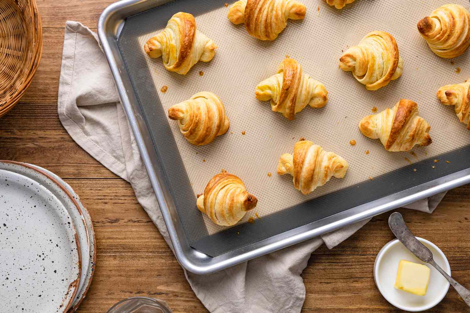 Homemade Crescent Rolls on a Baking Tray Surrounded by a Table Setting with Plates, a Small Saucer with Butter, and a Kitchen Towel
