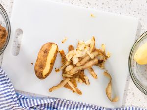 Potato peel and a half peeled potato on a white cutting board