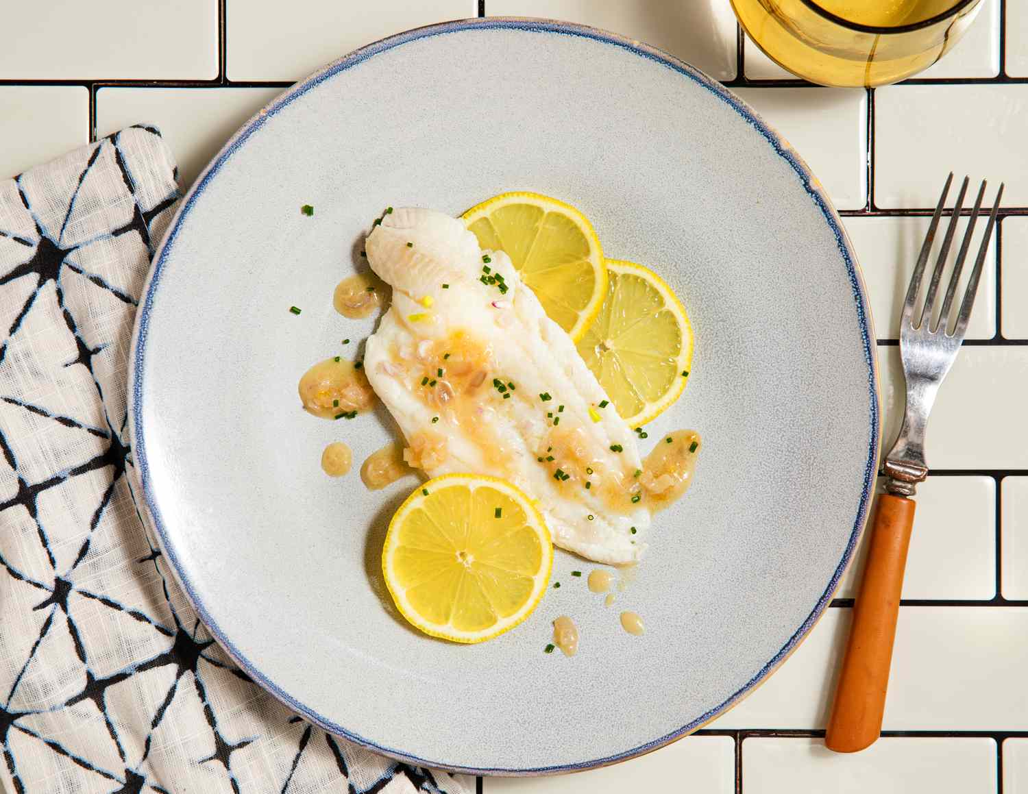 Prepared fish fillet on a plate, garnished with lemon slices and herbs, served on a white tile background with a napkin and fork nearby