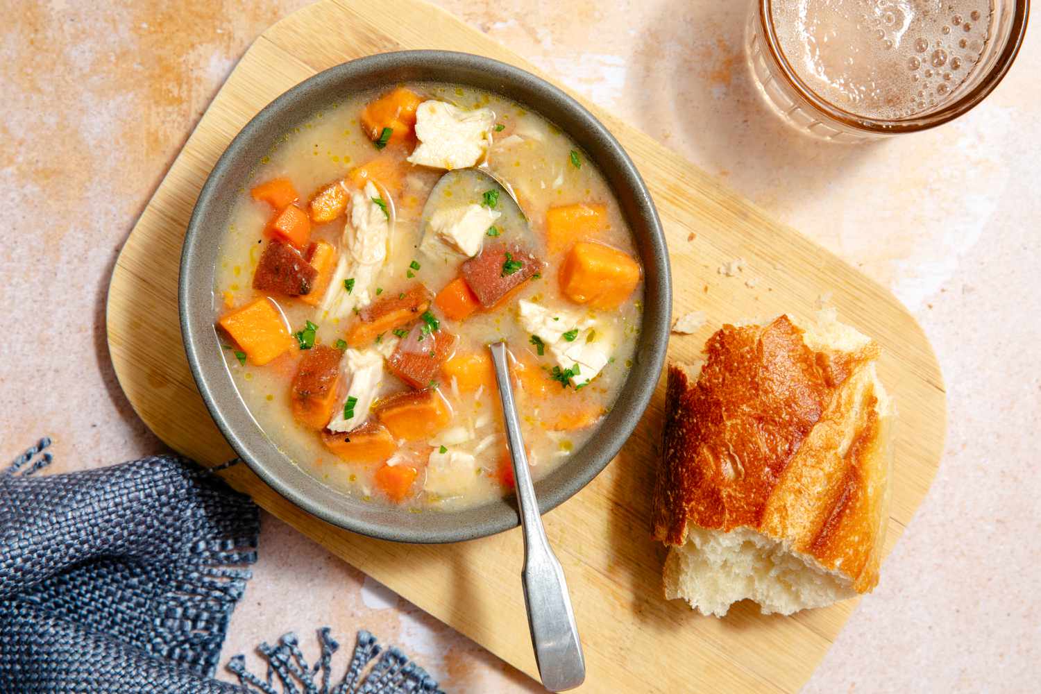 Overhead view of a gray bowl of chicken stew with a spoon all sitting on a wooden cutting board with a piece of french bread