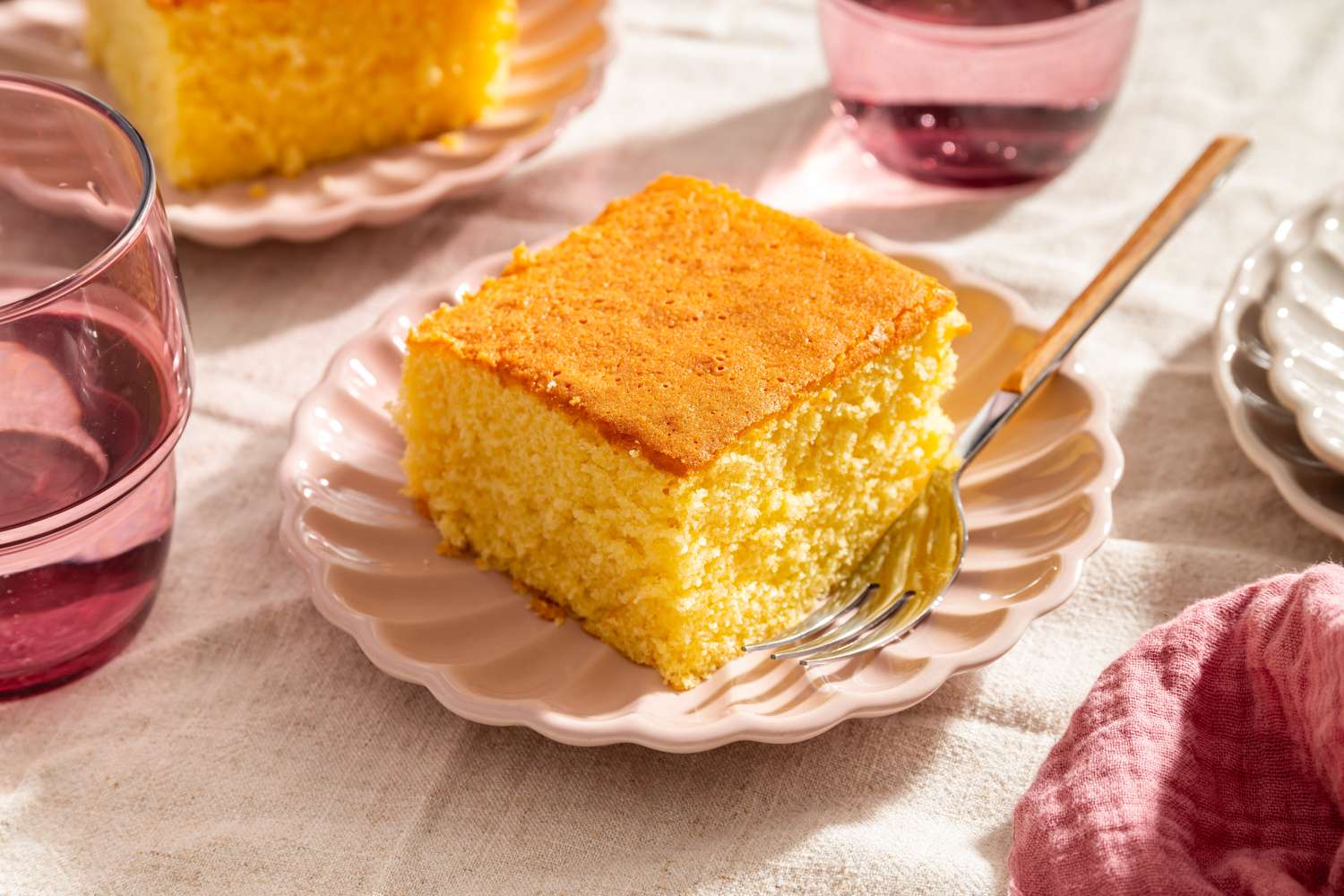A slice of hot milk cake on a scalloped plate with a fork surrounded by glasses and napkins on a table