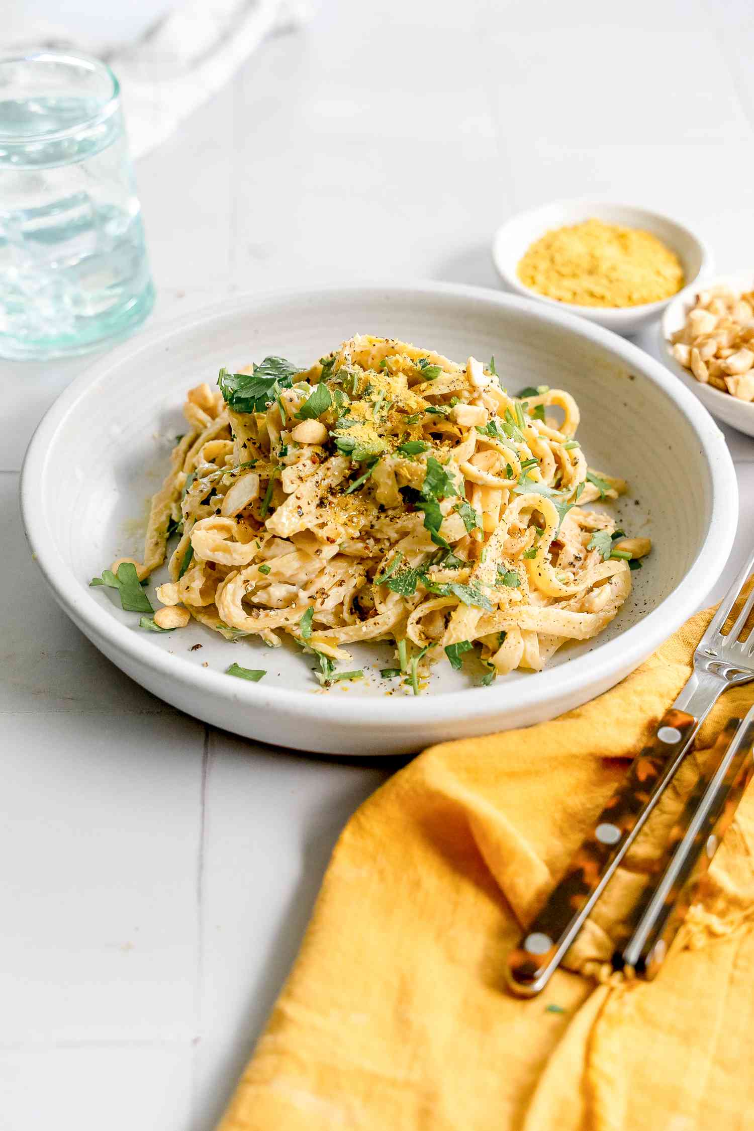 Plate of Super Creamy Vegan Pasta Topped with Parsley, and Next to It, Two Bowls of Ingredients (One: Cashews and Two: Nutritional Yeast), a Glass of Water, and a Utensils on a Table Napkin