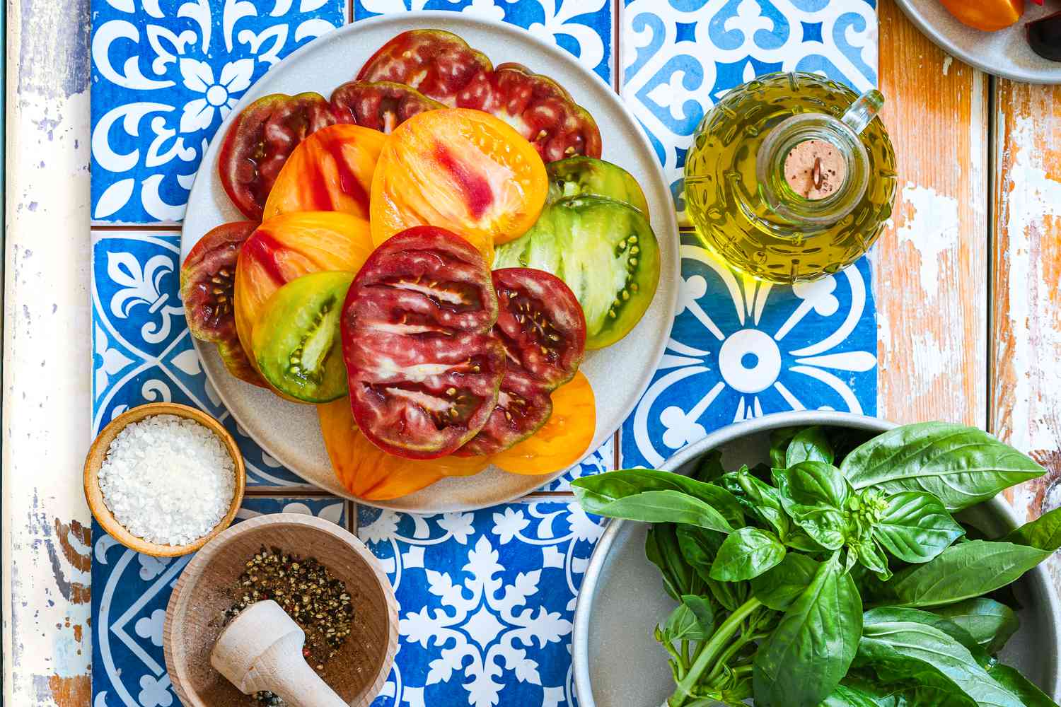 sliced heirloom tomatoes on a plate next to a bowl of coarse salt, crushed pepper in a mortar and pestle, carafe with olive oil, and a bowl of fresh basil, all on a spanish-tile covered tray