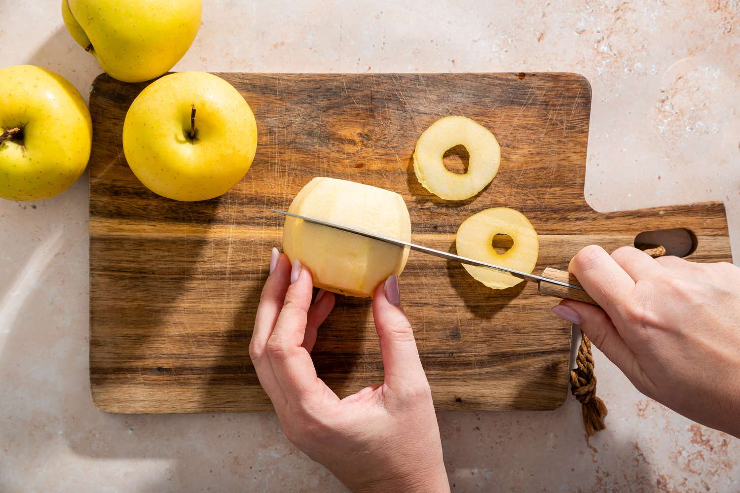 A person slicing an apple on a wooden cutting board beside whole apples and apple rings