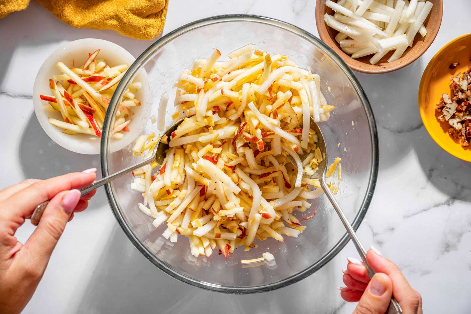 A bowl of kohlrabi apple salad being mixed with utensils with bowls of sliced ingredients around