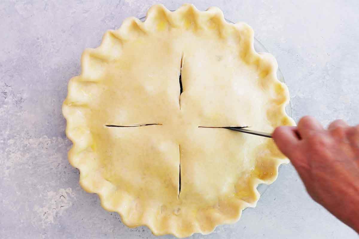 A knife cuts four slits into the top of the raw pie crust top 
