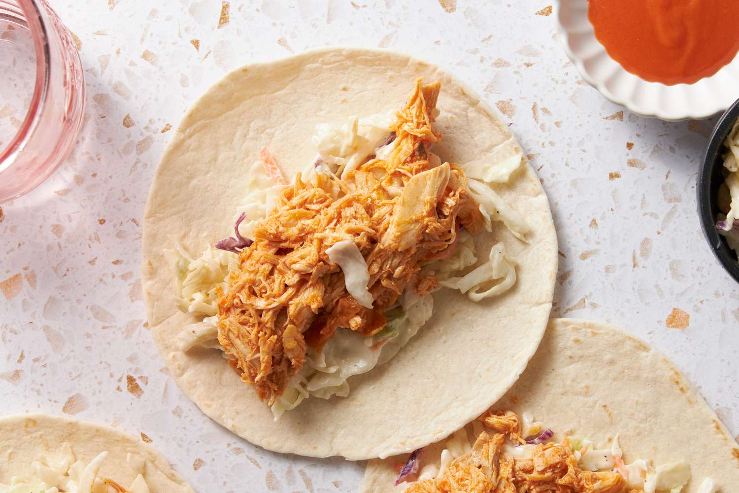 Buffalo Chicken and Coleslaw Added to Flour Tortilla, and in the Surroundings, More Tortillas With Filling, a Bowl With More Buffalo Sauce, a Bowl With Coleslaw, and a Glass