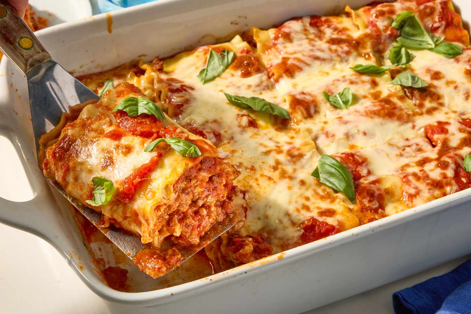 A casserole dish with lasagna rollups topped with melted cheese and basil one rollup being served