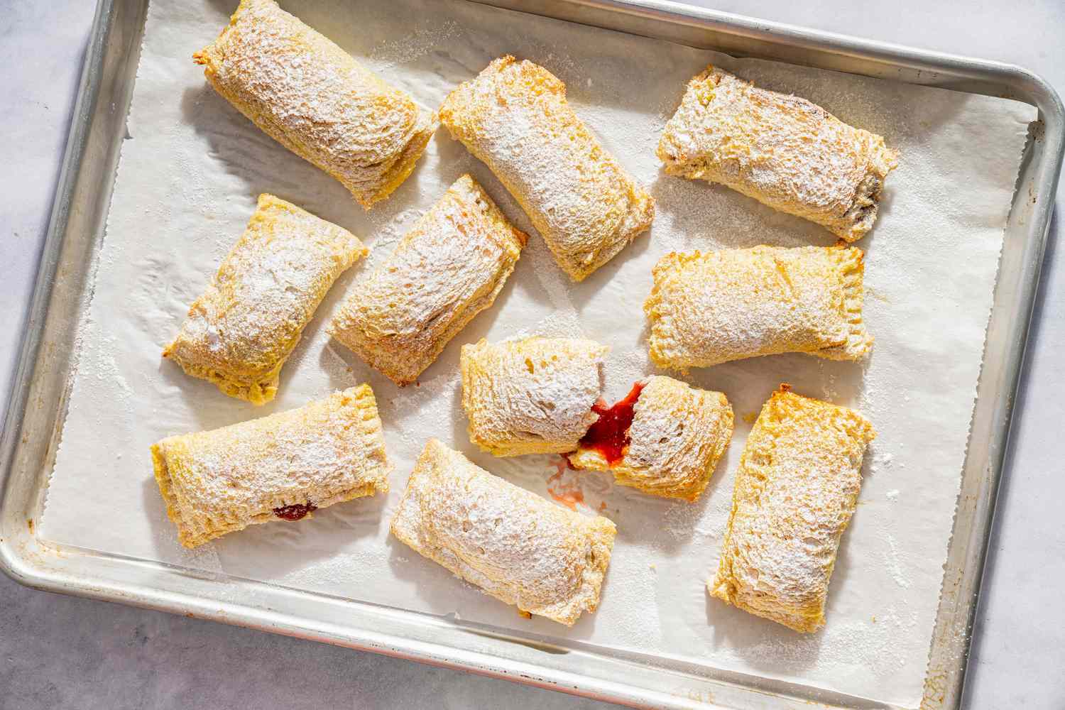 Toast Pies on a sheet-pan topped with parchment, dusted with powdered sugar, one torn open to show red filling