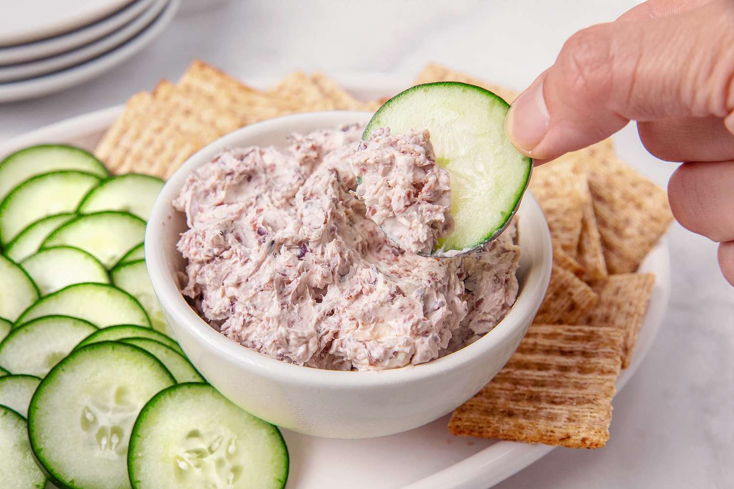 A bowl of cream cheese spread being scooped with a cucumber slice served with sliced cucumbers and crackers on a platter