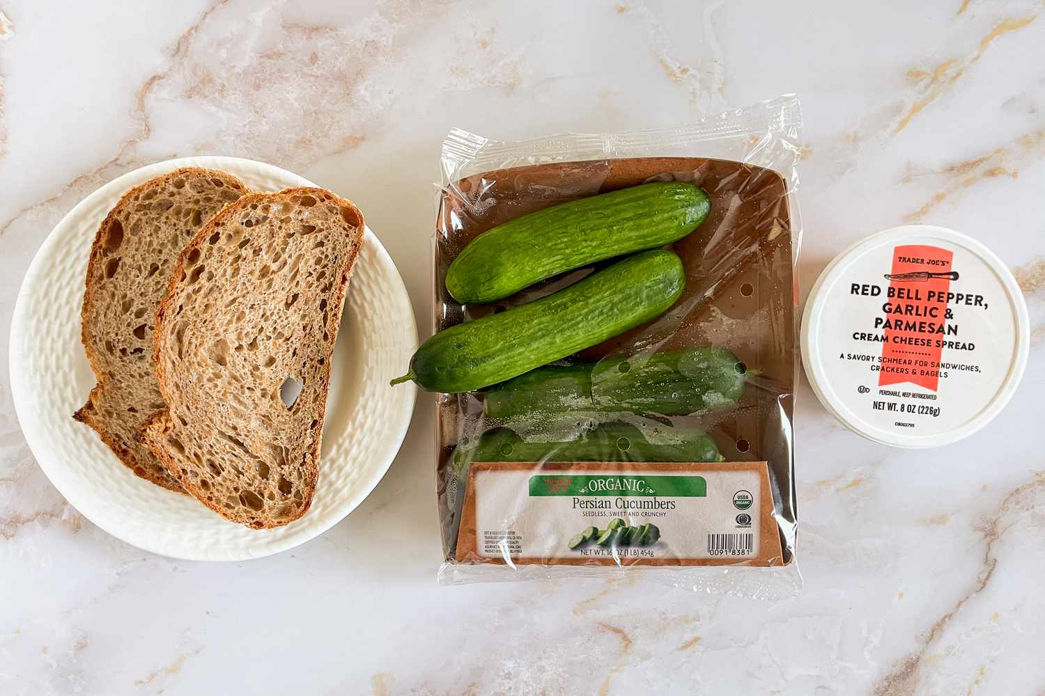 Ingredients for 2-ingredient cucumber sandwich (L to R): two slices of bread on a plate, a small package of Persian cucumbers, and a container of Trader Joe's Red Bell Pepper Garlic & Parmesan cream cheese