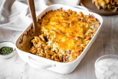 Casserole Dish of Cheeseburger Casserole with a Serving Already Served Using a Spoon at a Table Setting (a Plate with a Serving of Casserole, Bowl of Dry Parsley, a Table Napkin, and a Glass of Ice Water)