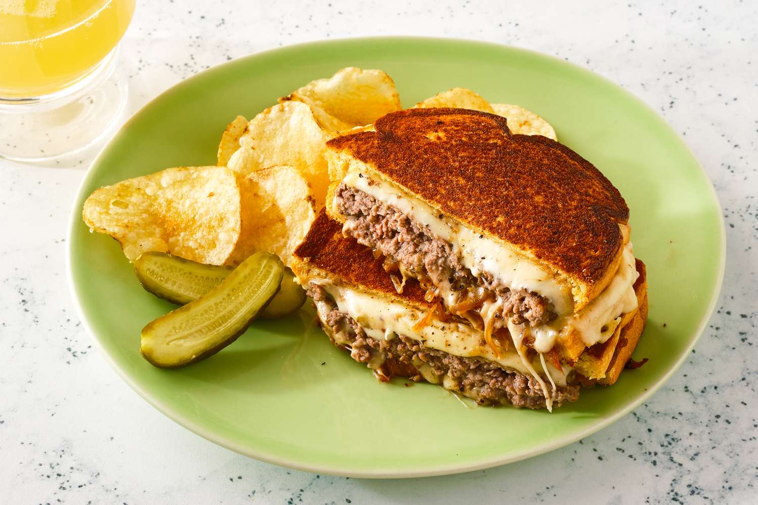 A patty melt, cut in half and stacked on a plate with chips and a pickle, ready to eat