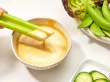 A stick of celery being dipped into a bowl of creamy mustard maple sauce, accompanied by fresh vegetables including snap peas and sliced cucumber