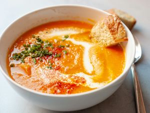 A bowl of carrot soup garnished with herbs and cream accompanied by a piece of bread