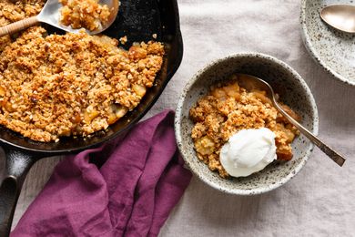Bowl of 3-ingredient skillet apple crisp with a dollop of yogurt next to a cast iron skillet with more apple crisp, a purple kitchen towel, and a bowl
