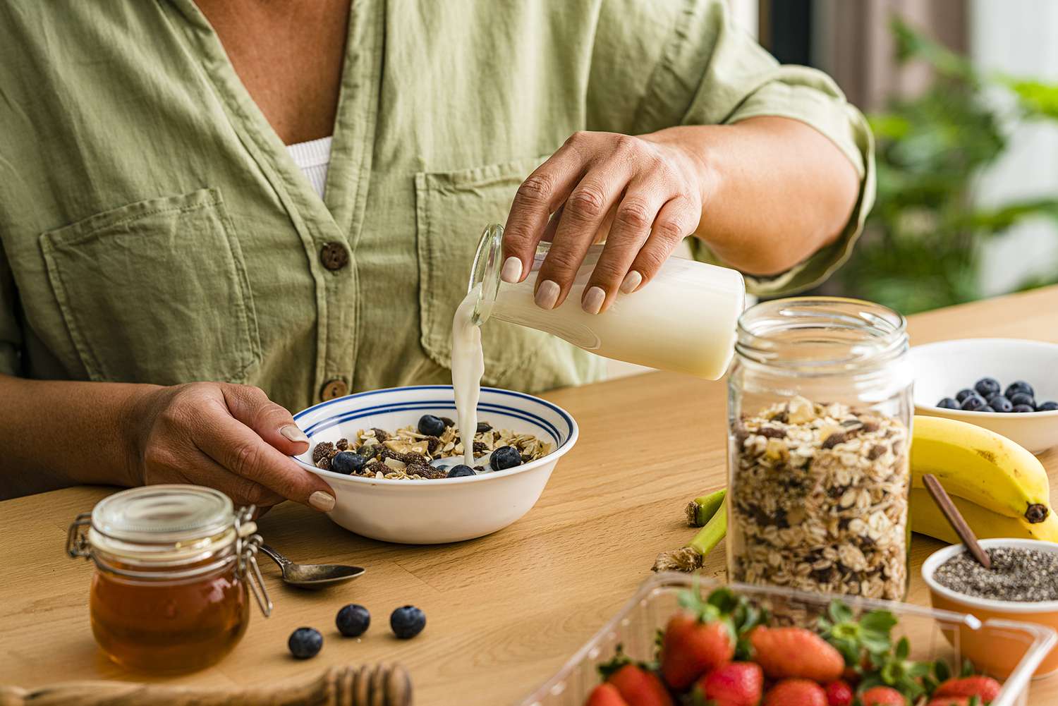 An angled view of a woman pouring almond milk into a bowl of granola at a table with a jar of honey, bowl of blueberries, container of strawberries and jar of granola