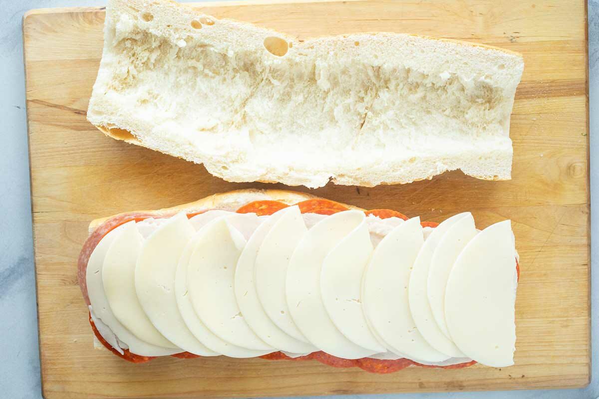 Showing how to make an italian sub sandwich by layering the bottom half with meat and cheese and scooping out the top of the bread.