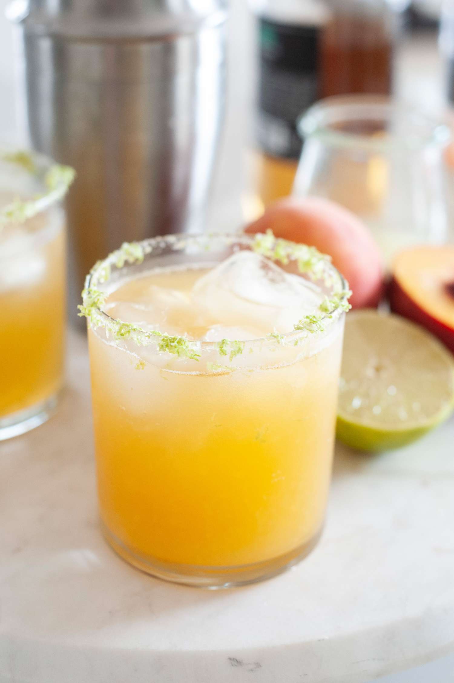 Classic margarita with peaches set on a tray with a cocktail shaker and halved peaches behind it.