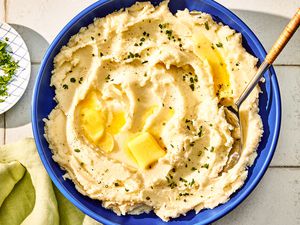 Overhead view of a large blue bowl of boursin mashed potatoes with a serving spoon and topped with herbs and butter all on a tile countertop