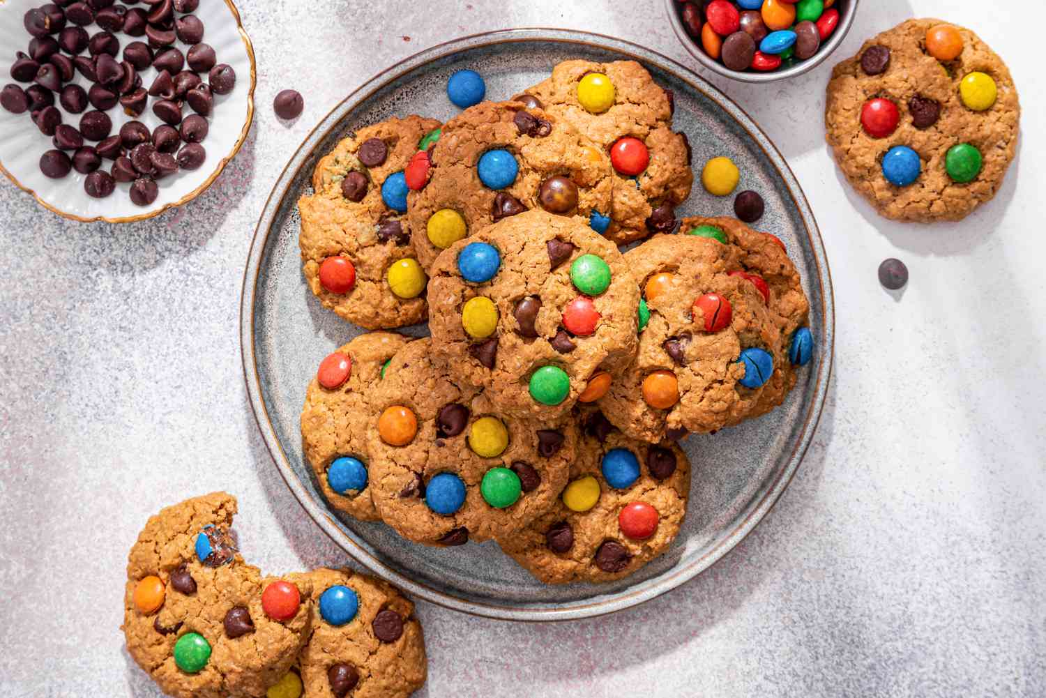 Plateful of monster cookies with some on the counter (a cookie with a bite missing also on the counter) next to a bowl of chocolate chips and a bowl of M&M's 
