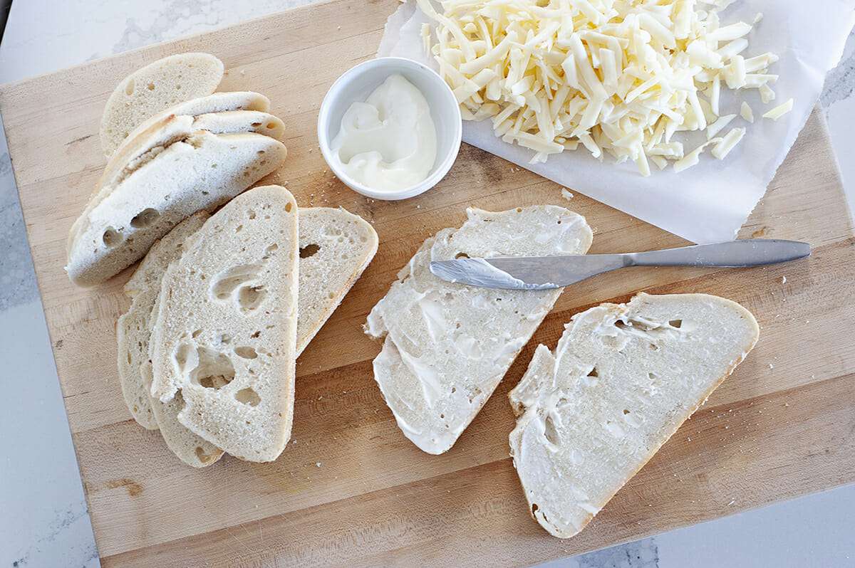 slices of bread spread with mayo, with a dish of mayo nearby and shredded cheese.