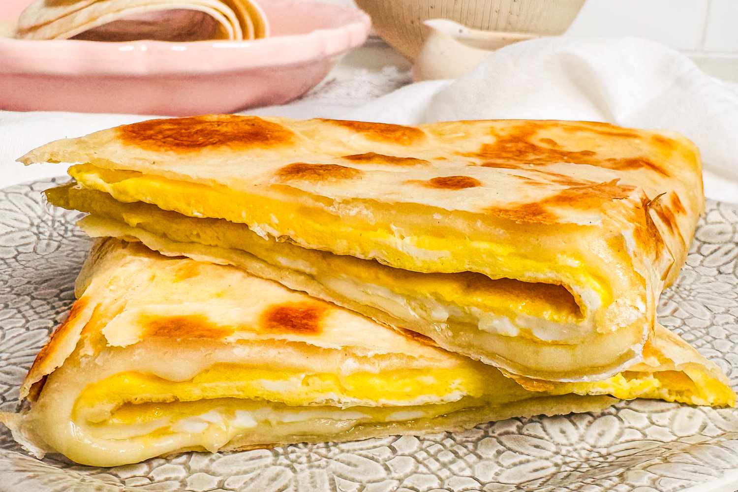 Folded egg tortilla on a patterned plate, layered with melted cheese, served with a dish in the background.
