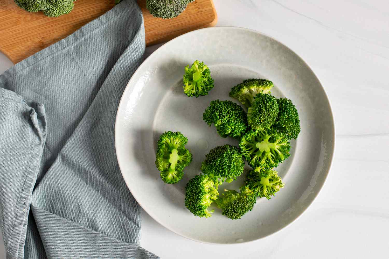 Blanched Broccoli on a Plate Surrounded by a Kitchen Towel and a Cutting Board with Heads of Broccoli for How to Blanch Broccoli
