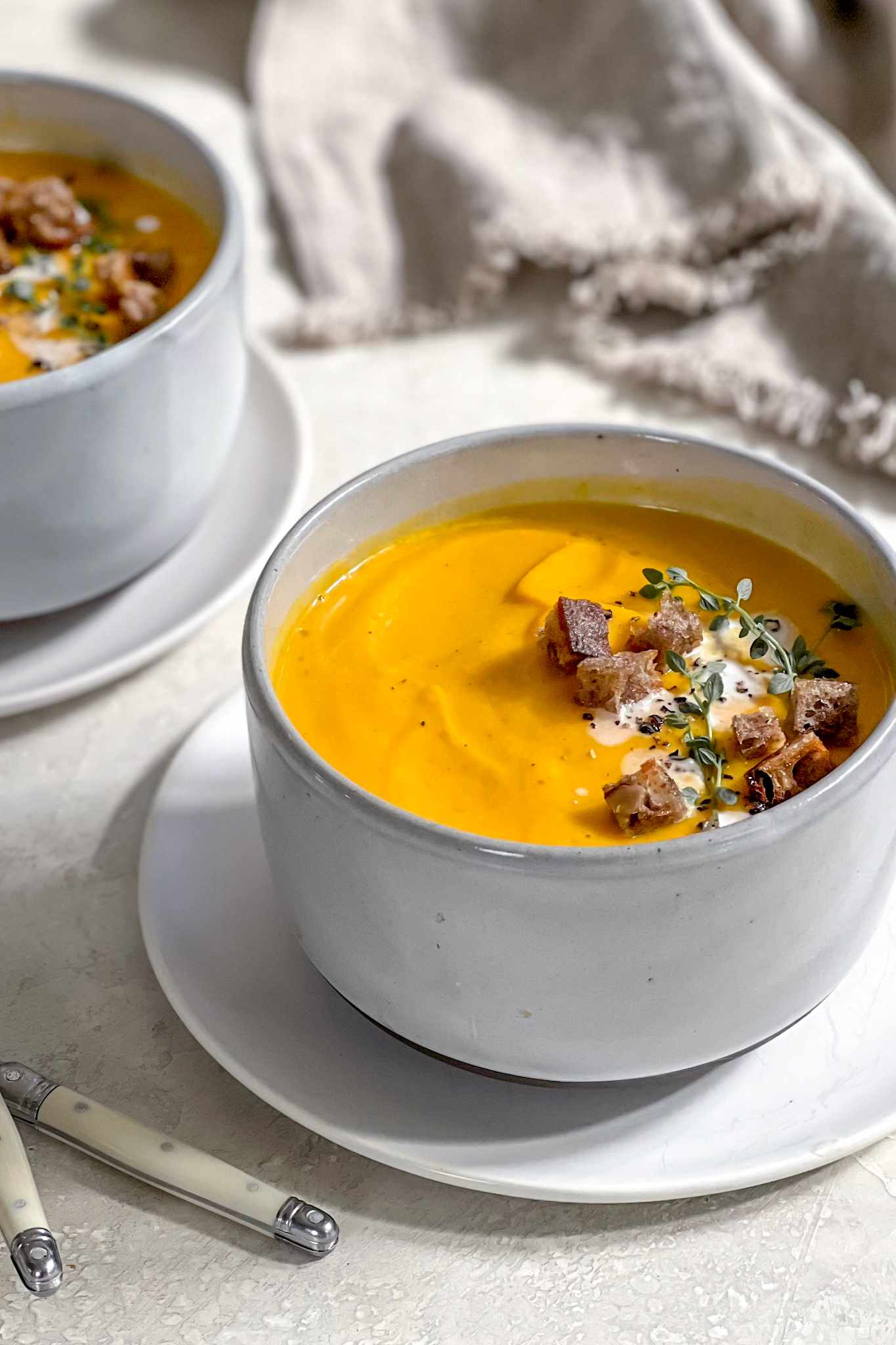 Two Bowls of Vegan Butternut Squash Soup Topped with Thyme, Coconut Milk, Croutons, and Cracked Pepper, Sitting on a Plate and in the Background, a Kitchen Linen