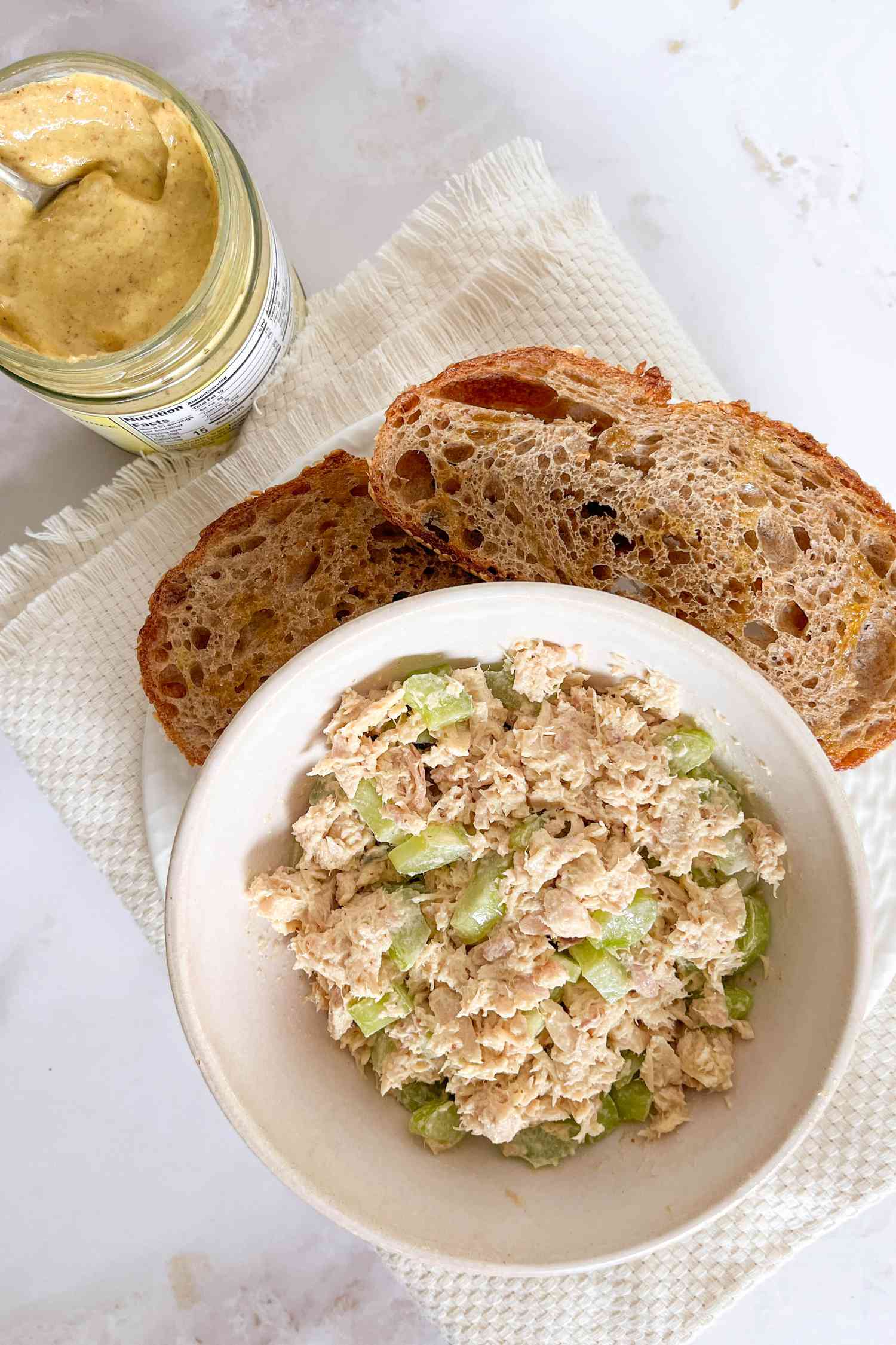 Tuna salad in a white bowl with bread on the side and a jar of mustard to the left
