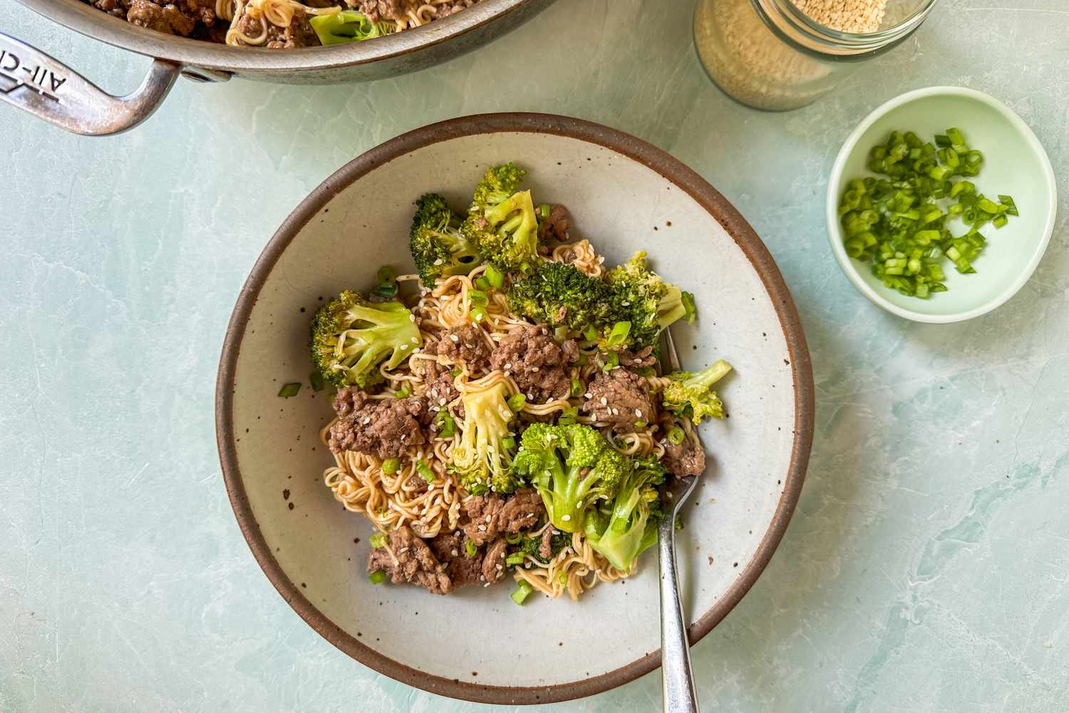 Overhead view of a brown rustic style bowl of ground beef, broccoli, noodles and a fork next to a small bowl of chopped green onions, a jar of sesame seeds on a light blue background