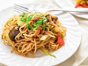 Side view of a white plate and fork with homemade vegetable lo mein. A fork is on the plate and a second plate is partially visible in the upper right hand corner.
