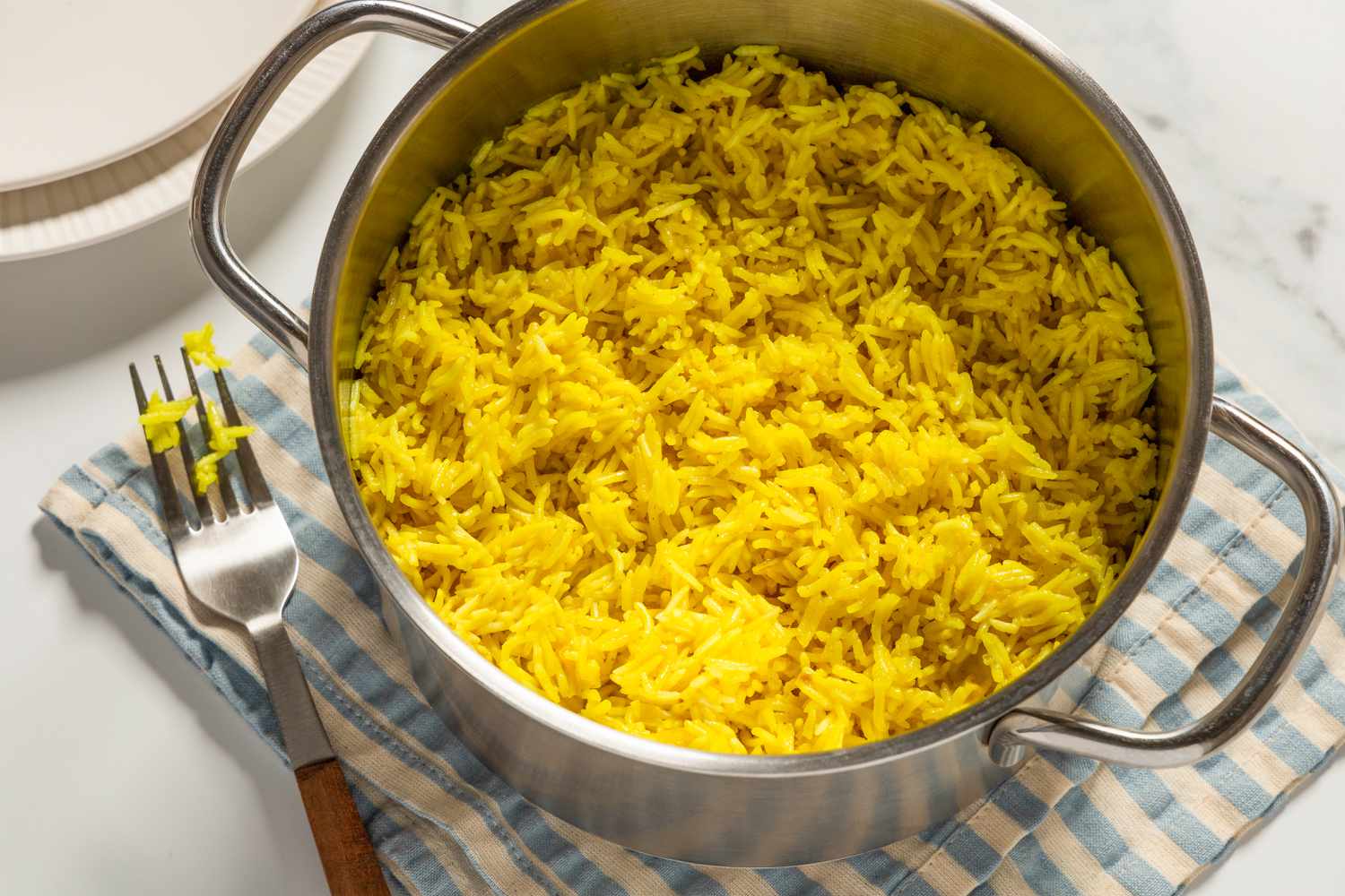 Overhead view of a pot of golden rice on marble countertop with a cloth napkin a fork and plates