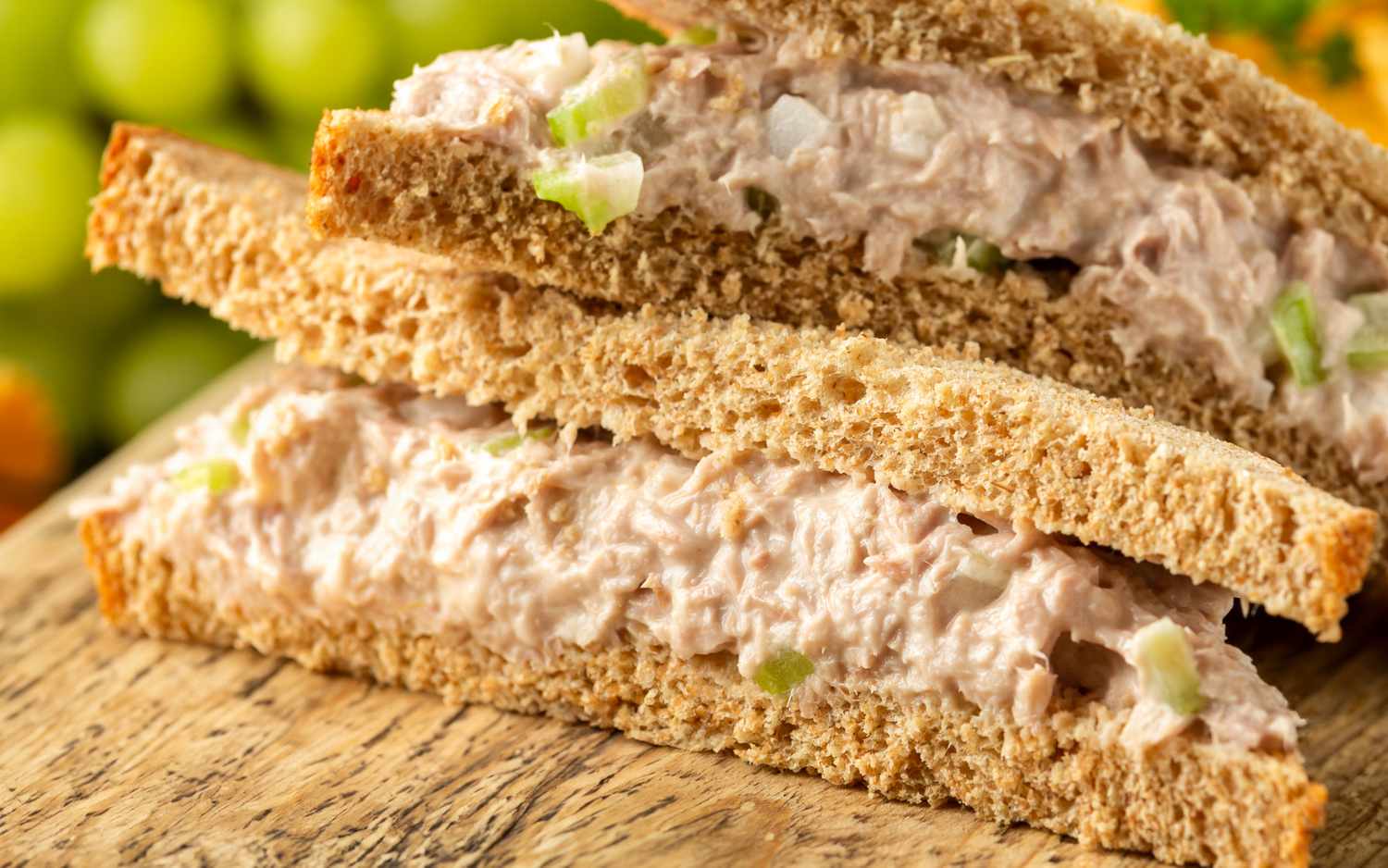 Two halves of a tuna salad sandwich on wholegrain bread placed on a wooden surface with blurry green grapes in background