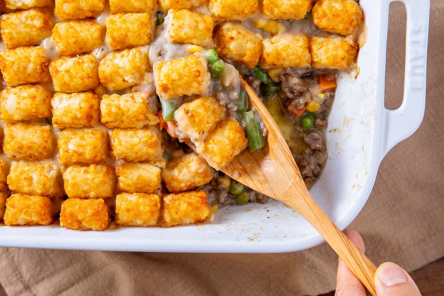 hand with wooden spoon scooping out some Classic Tater Tot Hotdish out of baking dish