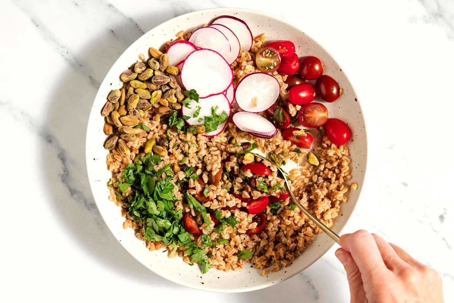 A farro salad with cherry tomatoes, radish slices, pistachios, and garnish, displayed in a bowl being stirred with a fork