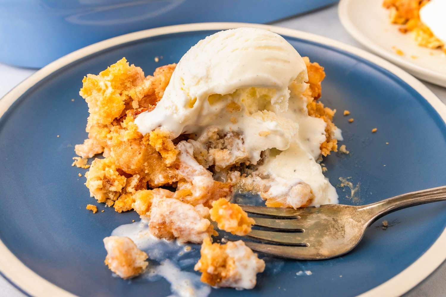 A closeup of a serving of apple dump cake topped with a scoop of vanilla ice cream, with a fork on the plate that has been cutting into the cake