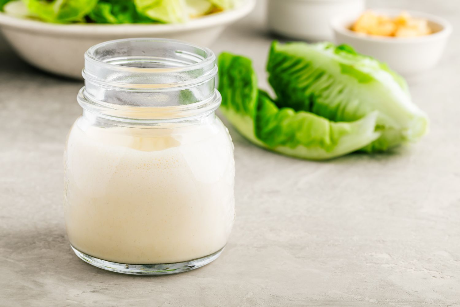 A glass jar of dressing on a countertop with lettuce in the background