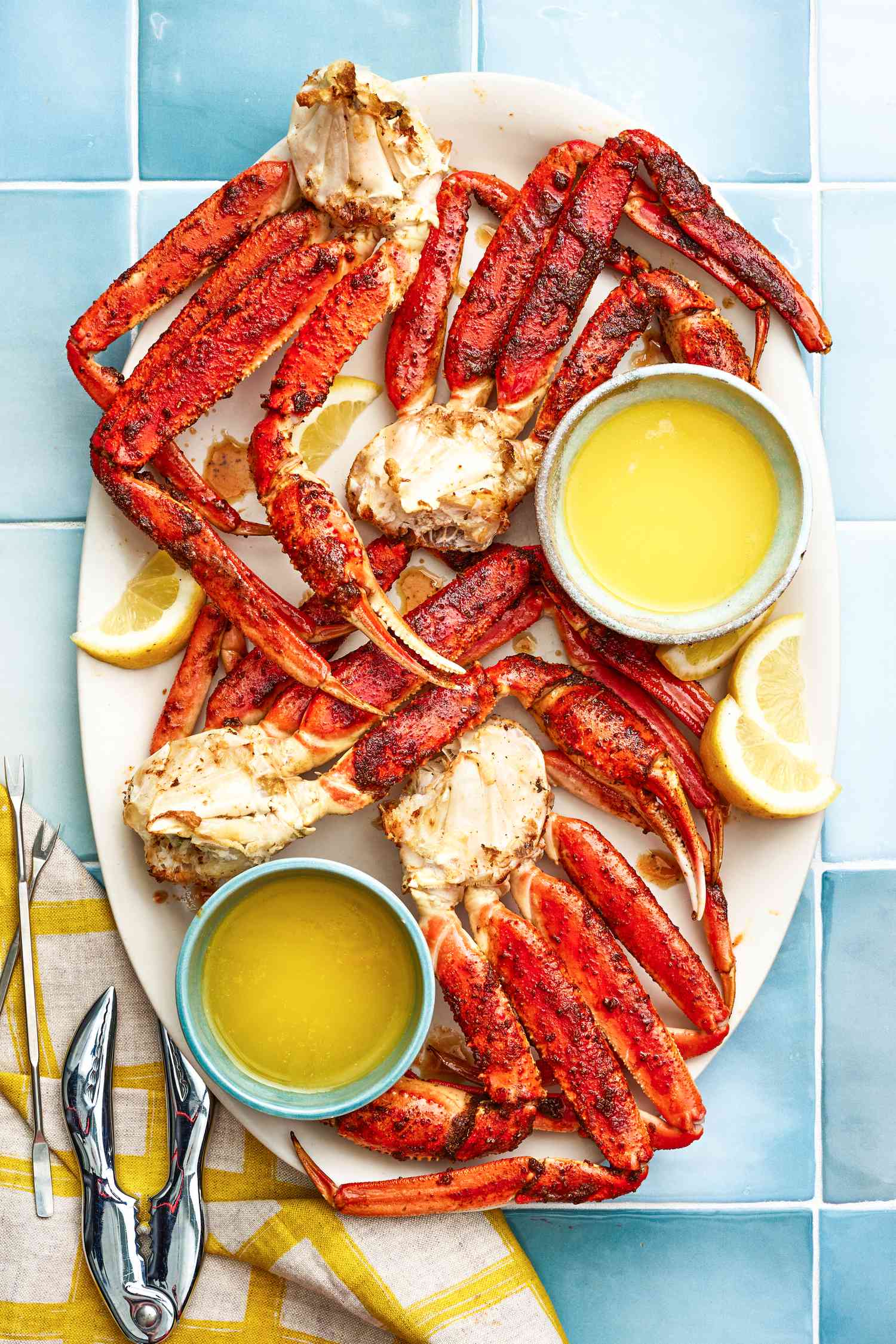 Serving Plate of Easy Baked King Crab Legs With Bowls of Melted Butter and Lemon Wedges, and on the Counter Next to It, a Few Seafood Picks and Cracker on a Table Napkin